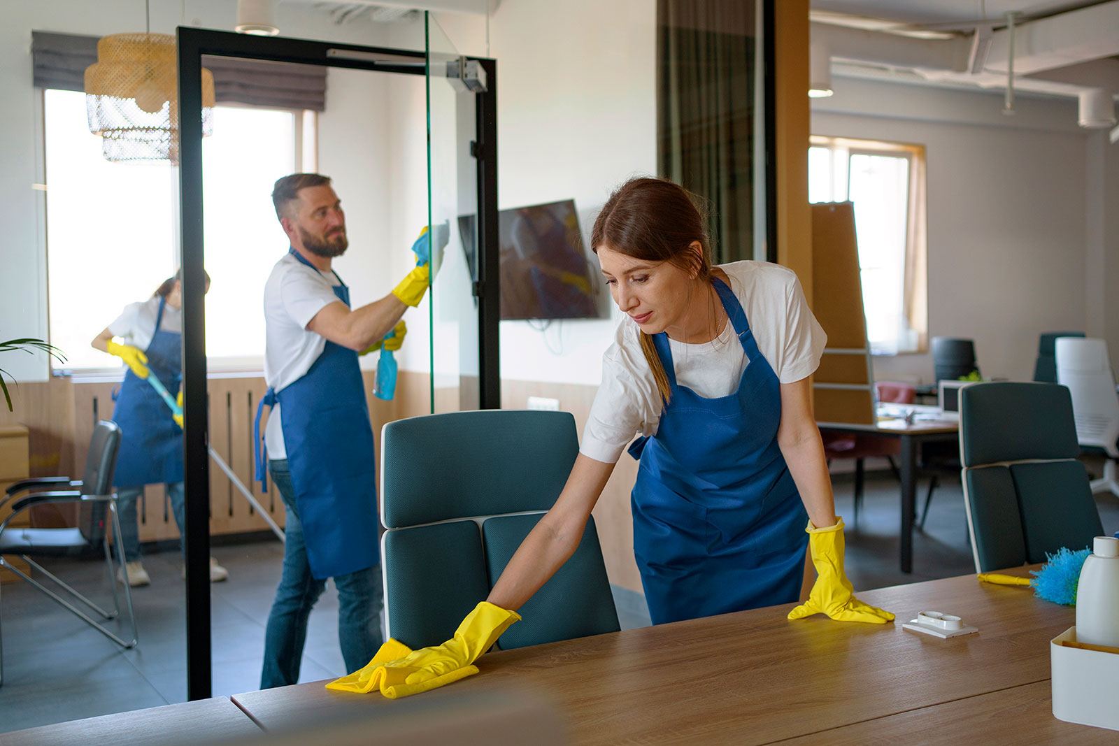 A woman wearing yellow cleaning gloves is wiping down a wooden desk in a bright, modern office space with large windows and plants in the background. She is focused on her task, and the room is well-lit by natural light.